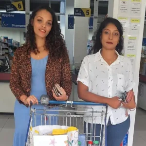 Two volunteers standing with a trolley filled with groceries and soft drinks at a store checkout.